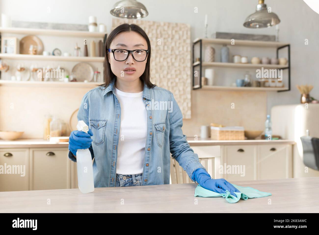 Portrait d'une jeune femme de ménage asiatique à la maison. Essuie la table dans la cuisine avec un chiffon dans des gants en caoutchouc, utilise des produits chimiques domestiques. Il est debout, tenant un spray, regardant la caméra. Banque D'Images