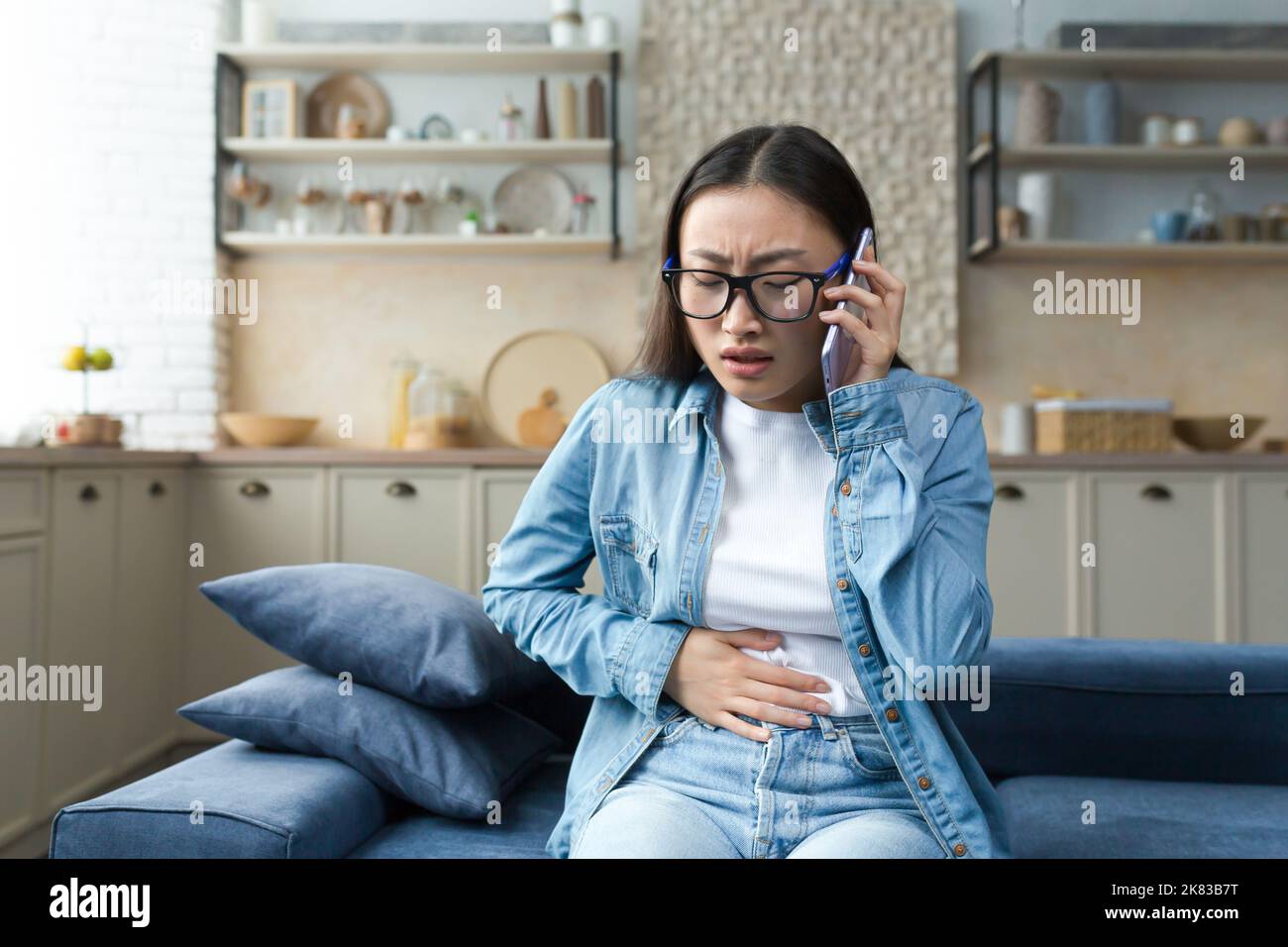 Une jeune femme asiatique enceinte, effrayée et anxieuse, assise sur le canapé à la maison et tenant son estomac, se sentant mal sévère, crampes dans son estomac. Il appelle une ambulance, appelle l'hôpital. Banque D'Images