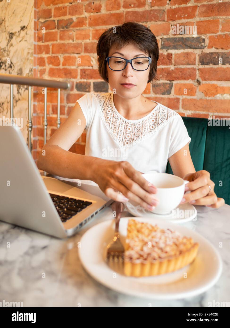 Femme travaille à distance dans un café avec des murs de briques rouges. Femme de type lunettes sur clavier d'ordinateur portable, boissons café et manger une tarte. Centre de coopération. Banque D'Images