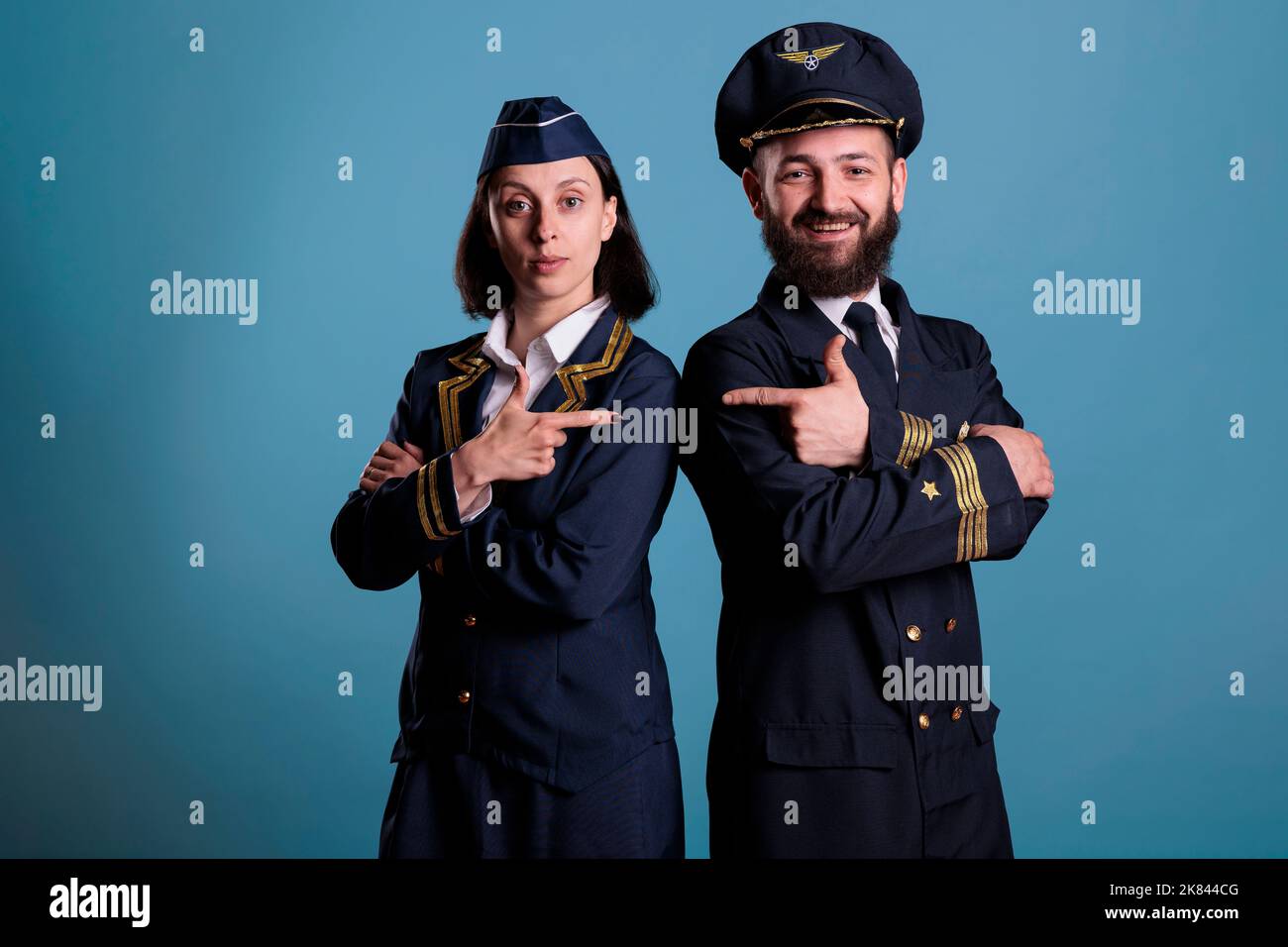 Portrait de l'avion capitan et de l'agent de vol se pointant l'un vers l'autre tout en étant debout en studio avec un fond bleu. Équipage d'aviation portant un uniforme professionnel, tir moyen Banque D'Images