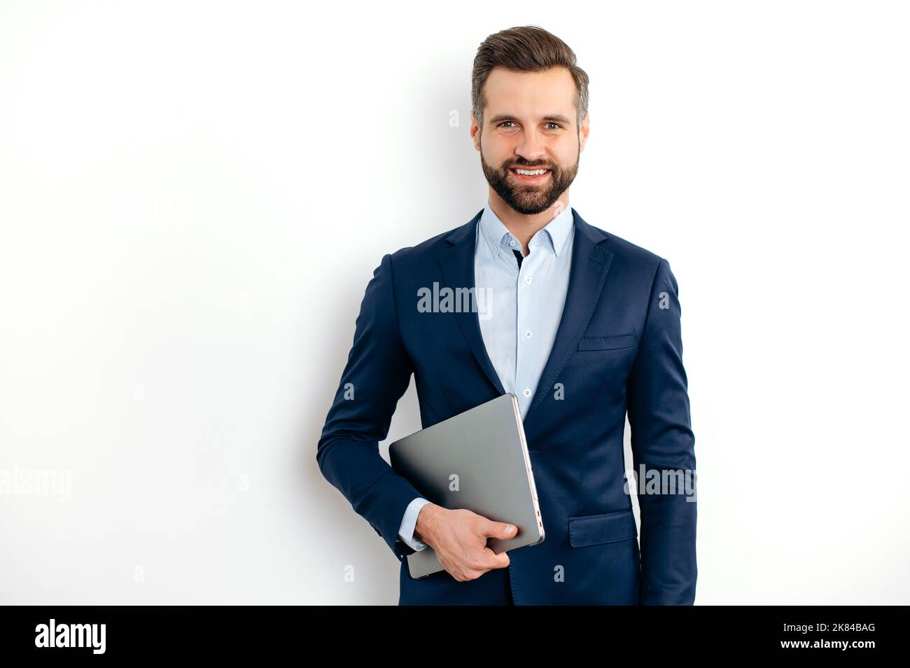 Portrait d'un homme d'affaires caucasien positif et réussi dans un costume, seo, consultant, courtier, debout sur un fond blanc isolé, tenant un ordinateur portable, regardant l'appareil photo, souriant amical Banque D'Images