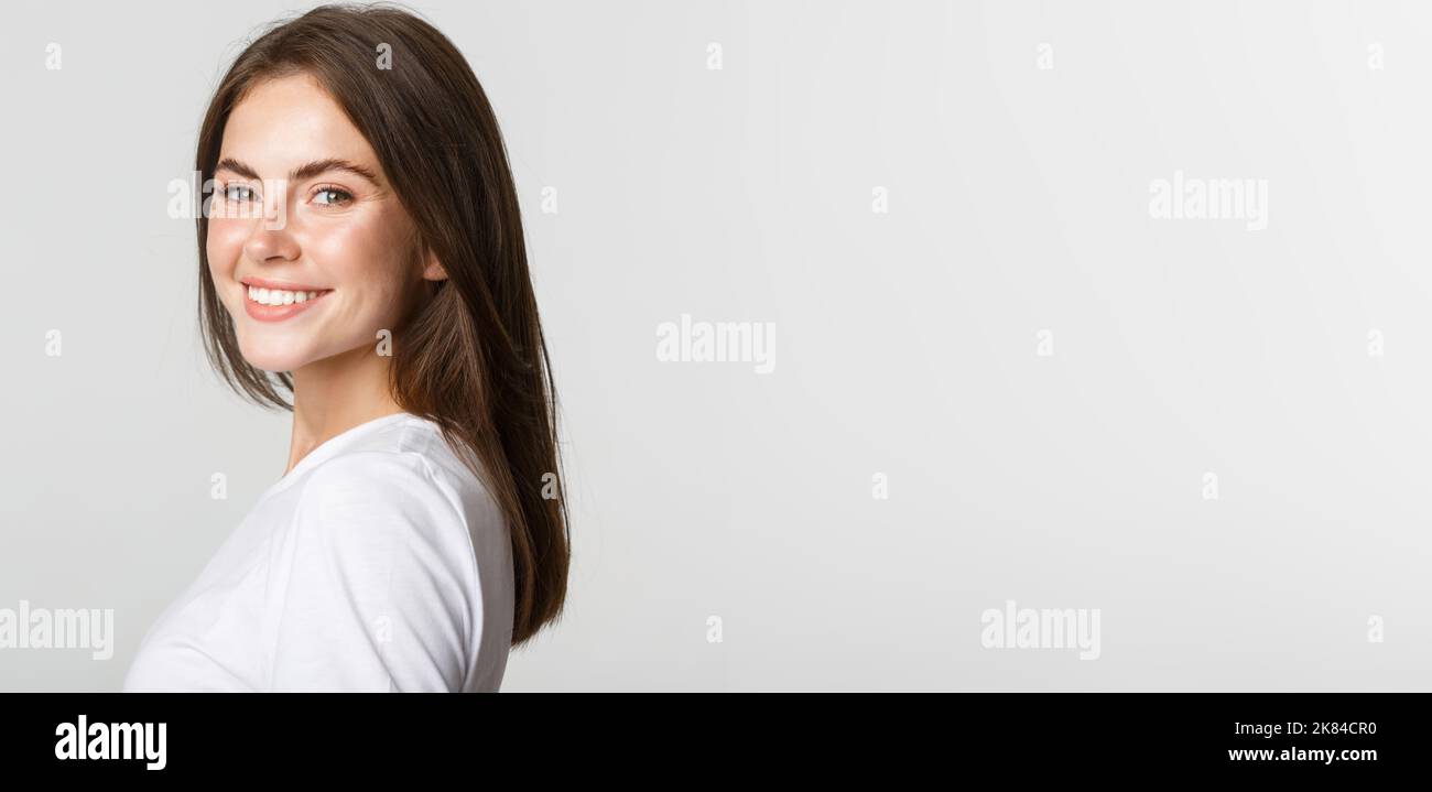 Portrait d'une femme brunette confiante qui tourne le visage de l'appareil photo avec un aspect rêveur, souriant sur fond blanc Banque D'Images