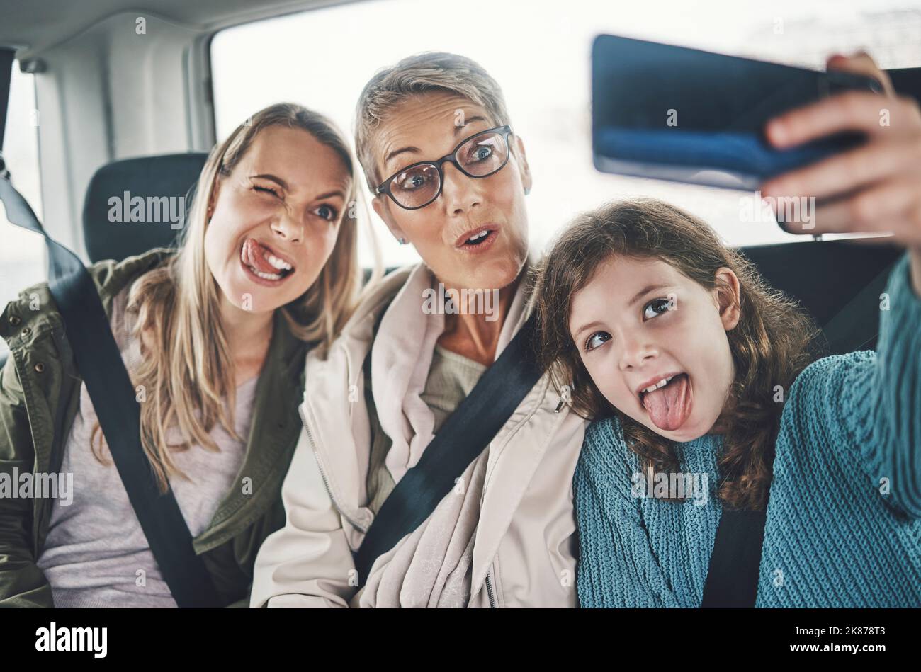 Téléphone, selfie et famille dans une voiture avec des visages stupides conduisant à une destination de vacances ensemble. Grand-mère, mère et fille enfant prenant la photo Banque D'Images