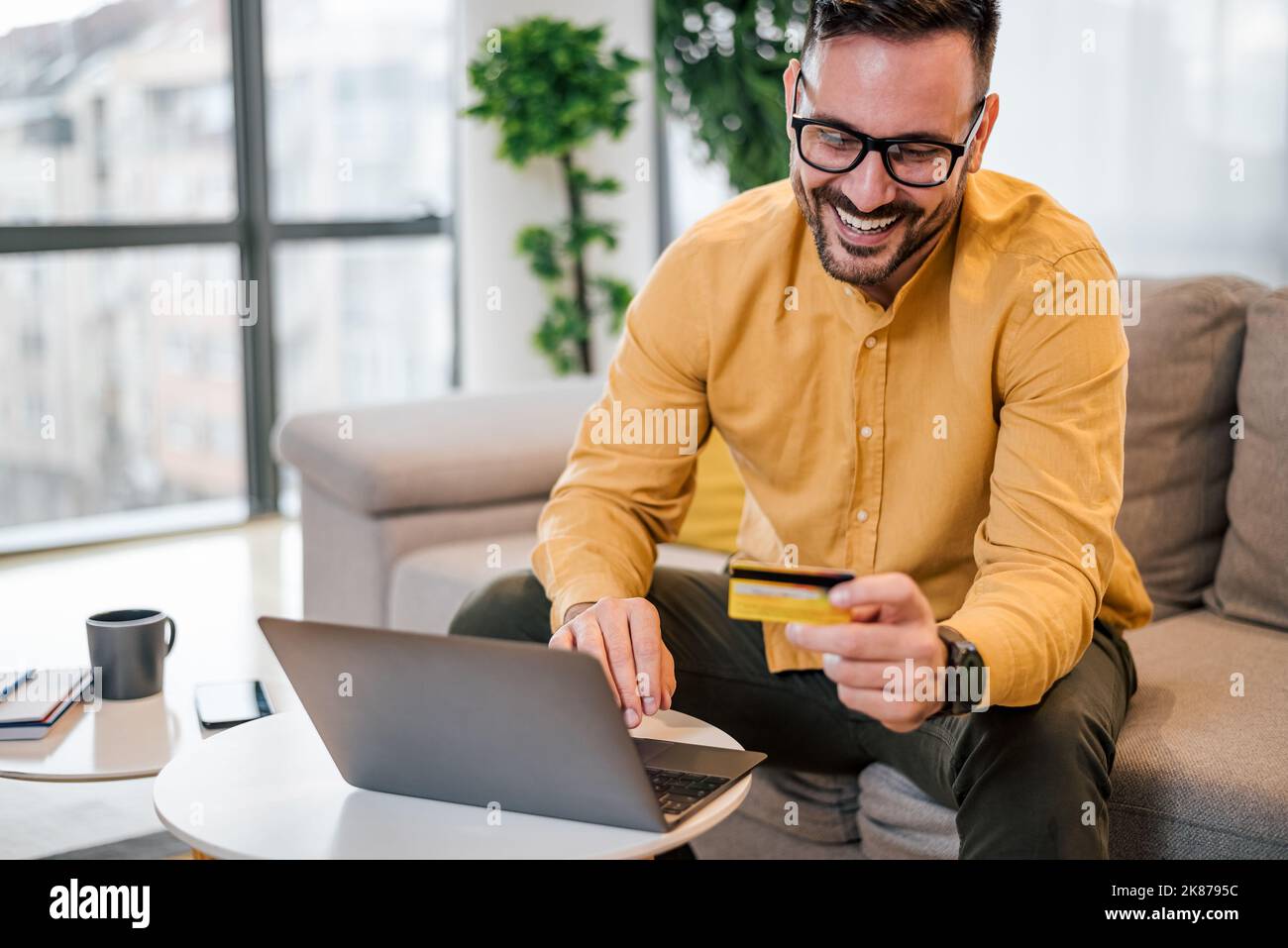 Ravi beau homme adulte avec des lunettes, souriant et lisant le numéro sur sa carte de crédit, à la maison. Banque D'Images
