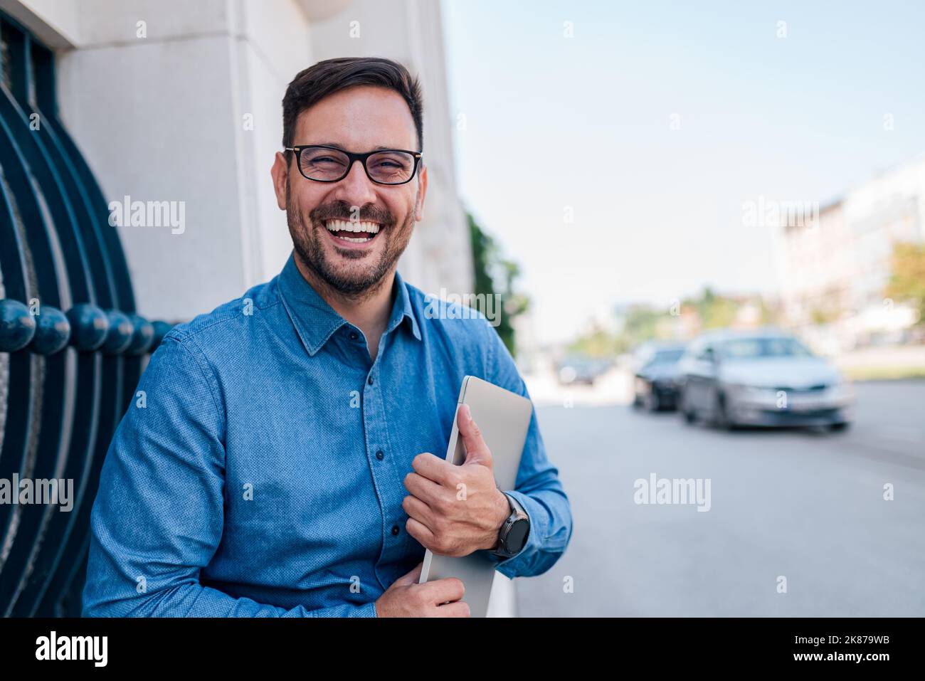 Photo ou bel homme adulte avec des lunettes, tenant fermement son ordinateur dans ses mains, souriant pour la photo. Banque D'Images