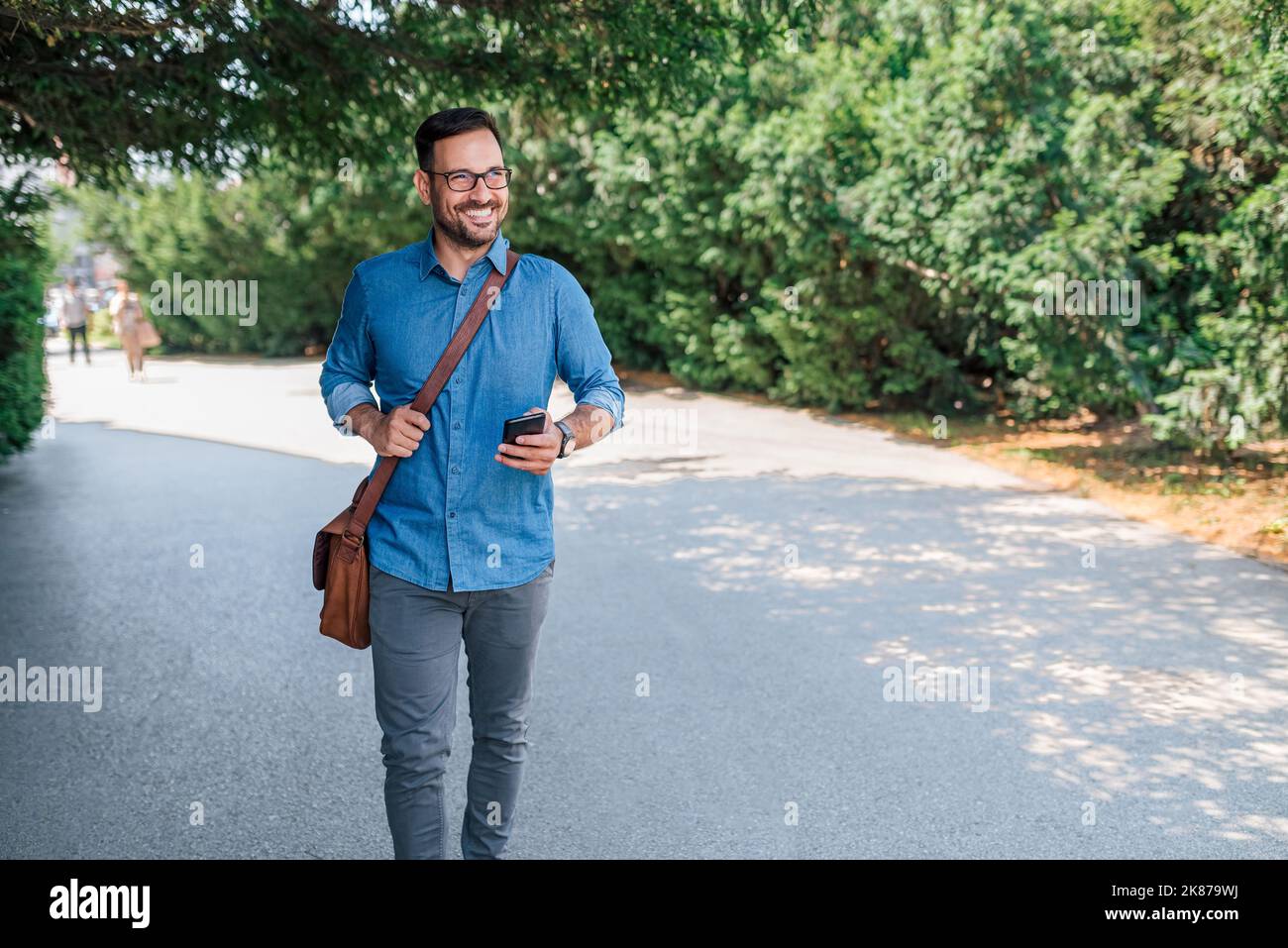 Homme barbu adulte souriant, portant son téléphone et son sac à main, profitant d'une belle journée à l'extérieur. Banque D'Images
