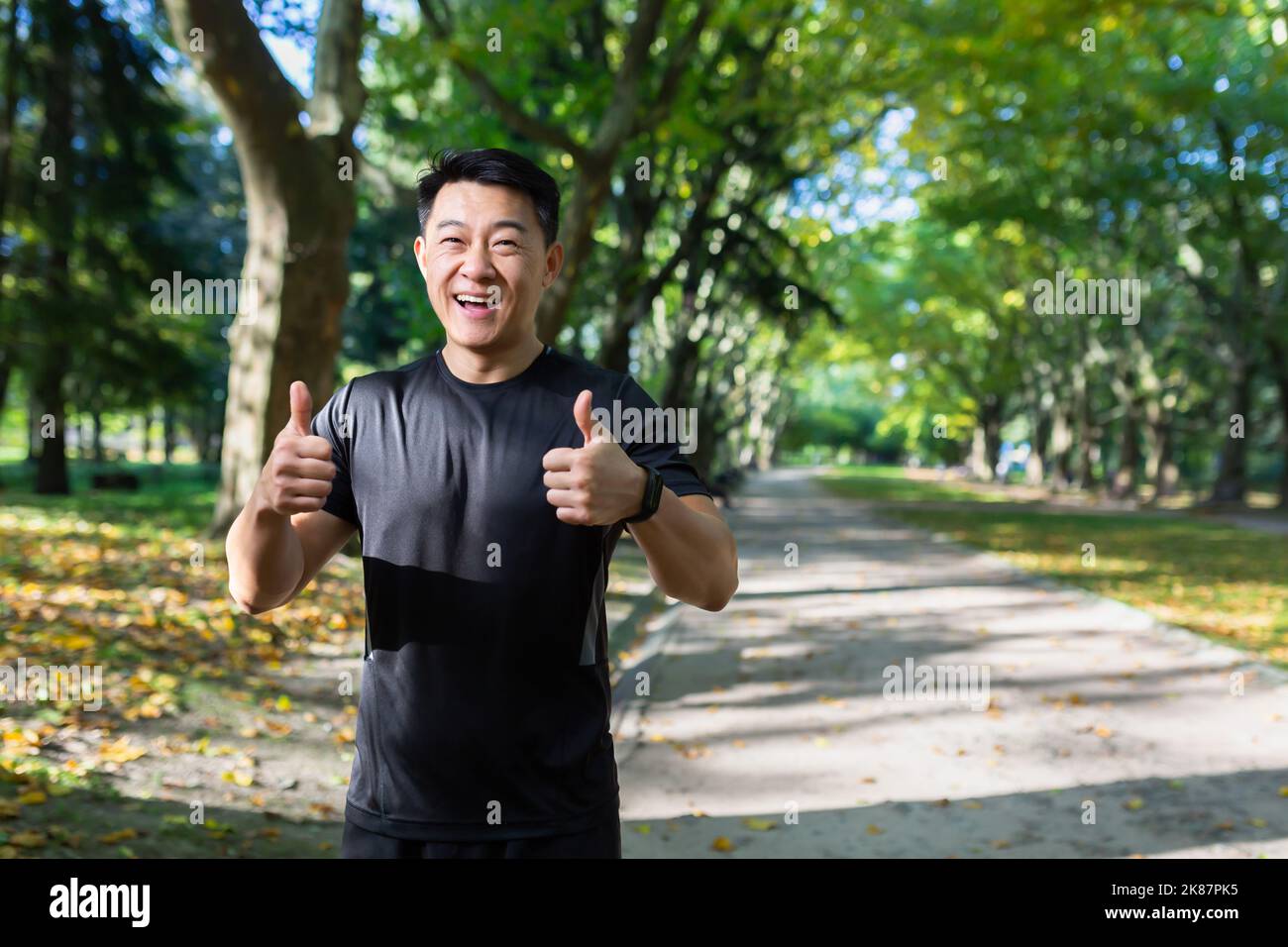 Porter de l'entraîneur de forme physique heureux dans le parc, homme asiatique souriant et regardant la caméra, pointant les doigts vers le haut affirmativement, homme faisant du sport en combinaison le jour ensoleillé près des arbres. Banque D'Images