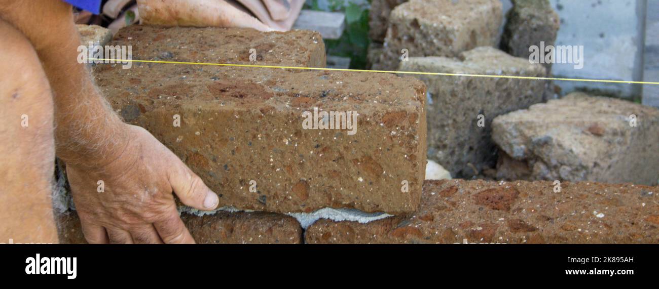 Images des mains d'un vieux maçon alors qu'avec du ciment il construit un mur bas avec des briques tuff. Homme au travail. Bannière horizontale Banque D'Images