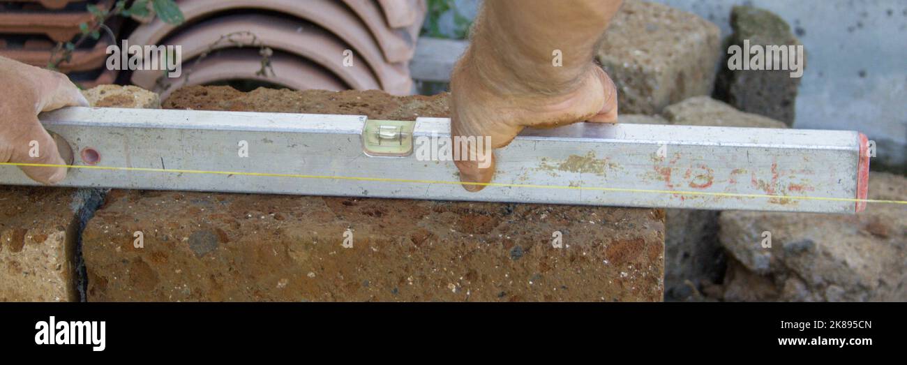 Image des mains d'un bricklayer qui vérifie la régularité du plan du mur de briques tuf qu'il construit avec un niveau. Bannière horizontale Banque D'Images