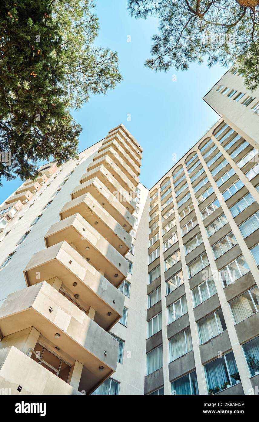 Ancien bâtiment urbain de haute élévation soviétique avec balcons et fenêtres voûtées contre de végétation luxuriante et ciel clair par jour ensoleillé dans la ville vue à angle bas. Banque D'Images