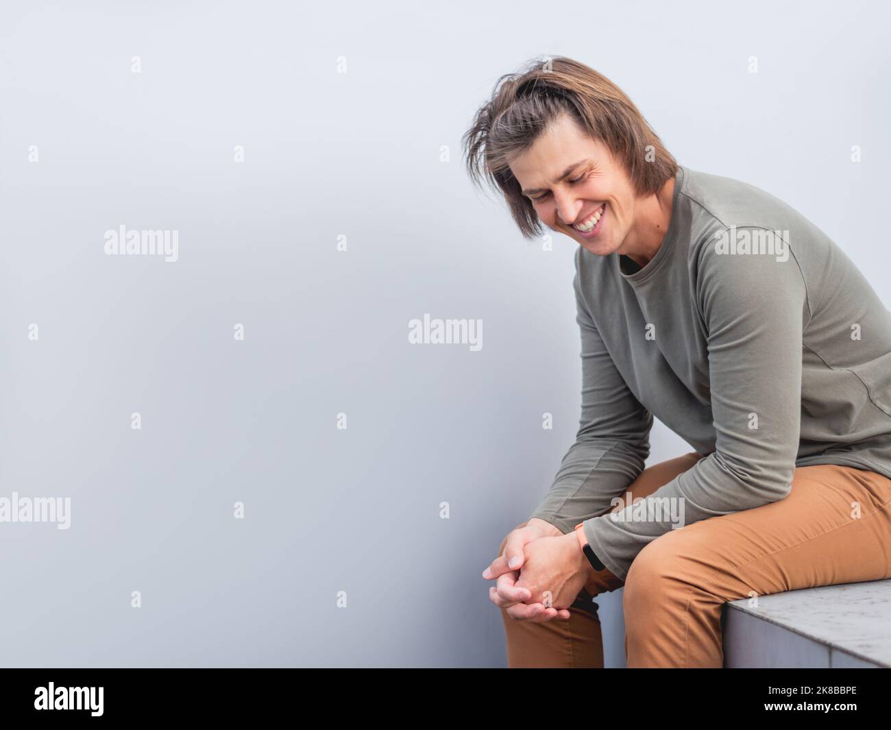 Homme aux cheveux longs et au look heureux se trouve près du mur gris. Homme souriant dans des vêtements décontractés de terre rient sur la bannière horizontale avec l'espace de copie. Banque D'Images