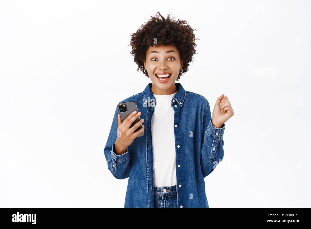 Une jeune femme heureuse et belle saute de l'excitation, en utilisant un téléphone mobile et en regardant joyeusement, en gagnant sur l'application pour smartphone, debout sur fond blanc Banque D'Images