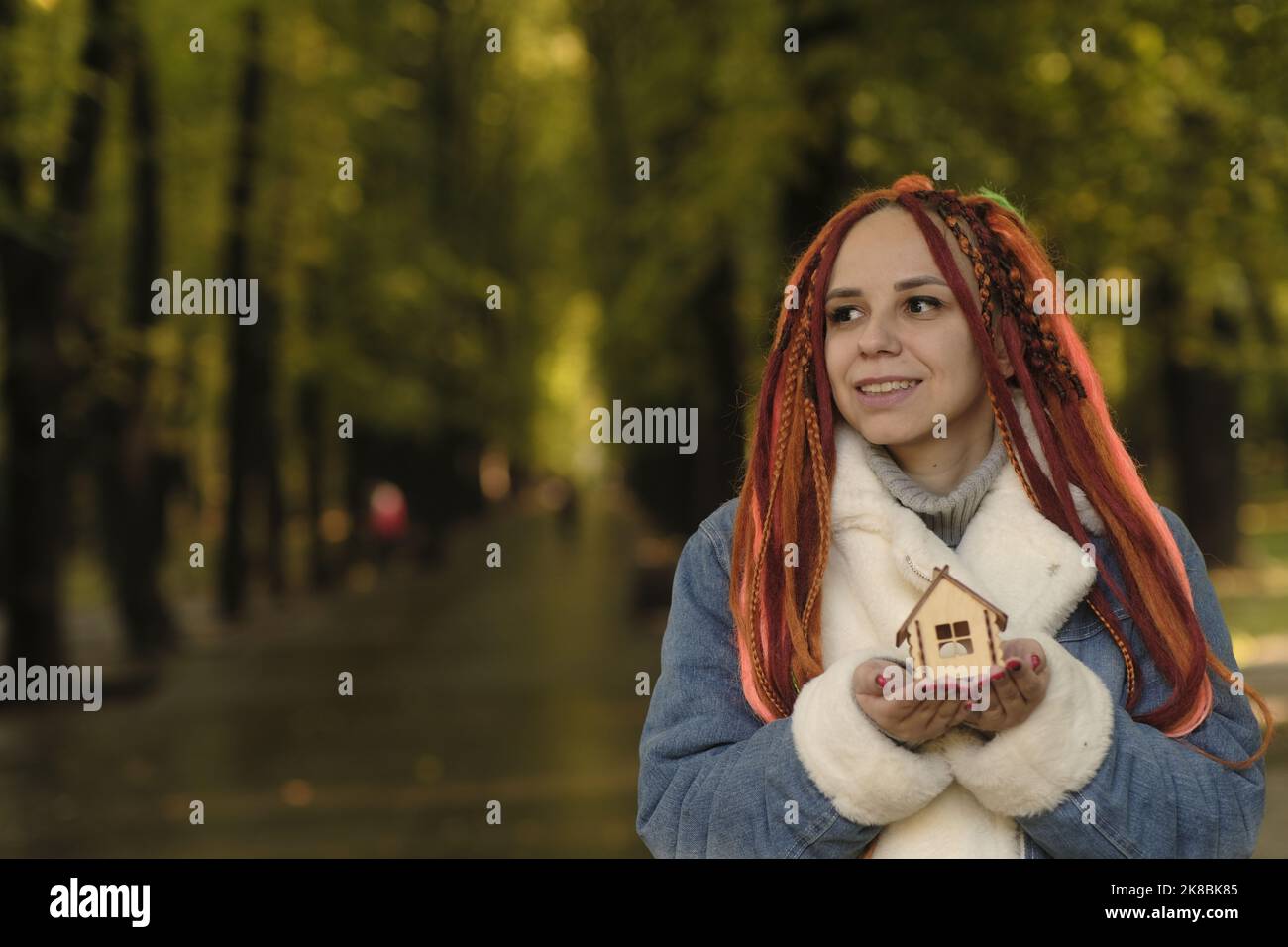 Bonne femme tenant le modèle à la maison dans le parc. Jeune femme positive avec des dreadlocks tenant une petite maison en bois souriant et regardant la caméra dans le parc d'automne. Banque D'Images