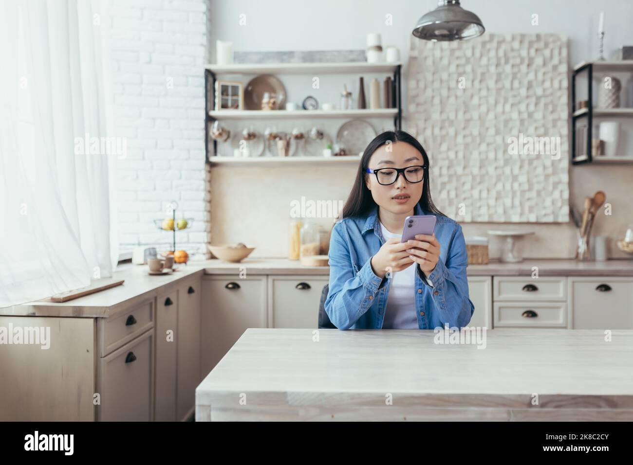 Jeune femme asiatique en lunettes et vêtements en denim, assise dans la cuisine à la maison et à l'aide d'un téléphone portable. Cadrans, sourires. Banque D'Images
