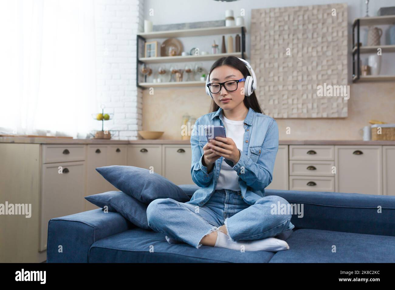Une jeune fille étudiante asiatique est assise sur le canapé à la maison en position lotus. Il écoute un podcast, un livre audio, étudie dans un casque blanc avec un téléphone entre ses mains. Banque D'Images