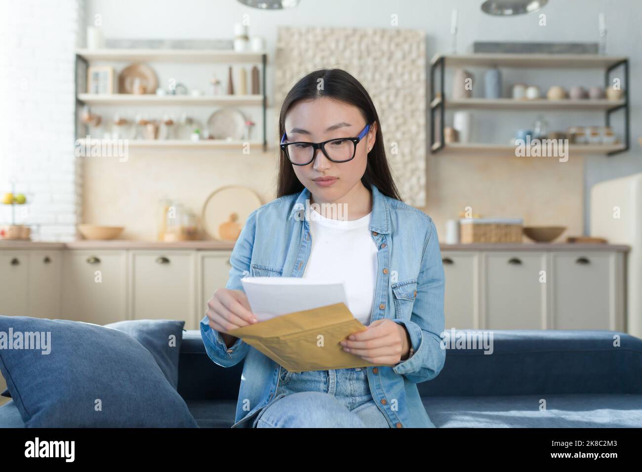 Une jeune fille étudiante asiatique magnifique a reçu une lettre d'admission à l'université dans le courrier. Elle tient une enveloppe entre ses mains. Assis à la maison sur le canapé, lecture. Banque D'Images