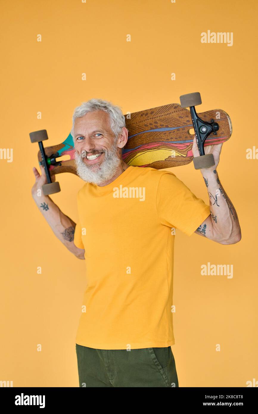 Patineur homme senior à barbe heureux tenant un skateboard isolé sur jaune. Banque D'Images