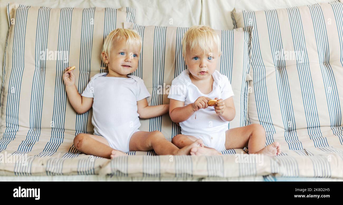 Adorables enfants identiques avec la lumière entendre et les yeux bleu ciel. Les jumeaux de bébé blancs s'assoient sur un confortable canapé beige dans le salon Banque D'Images