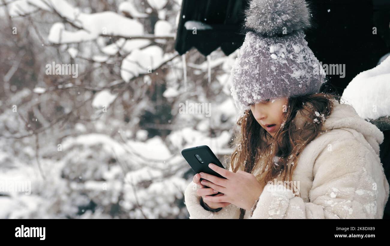 portrait d'hiver. cutie petite fille, vêtue de vêtements chauds d'hiver, entièrement couverte de flocons de neige, est assise sur banc et à l'aide d'un smartphone, à l'extérieur, en chute de neige, près de la vieille maison en bois. jour d'hiver enneigé. Photo de haute qualité Banque D'Images