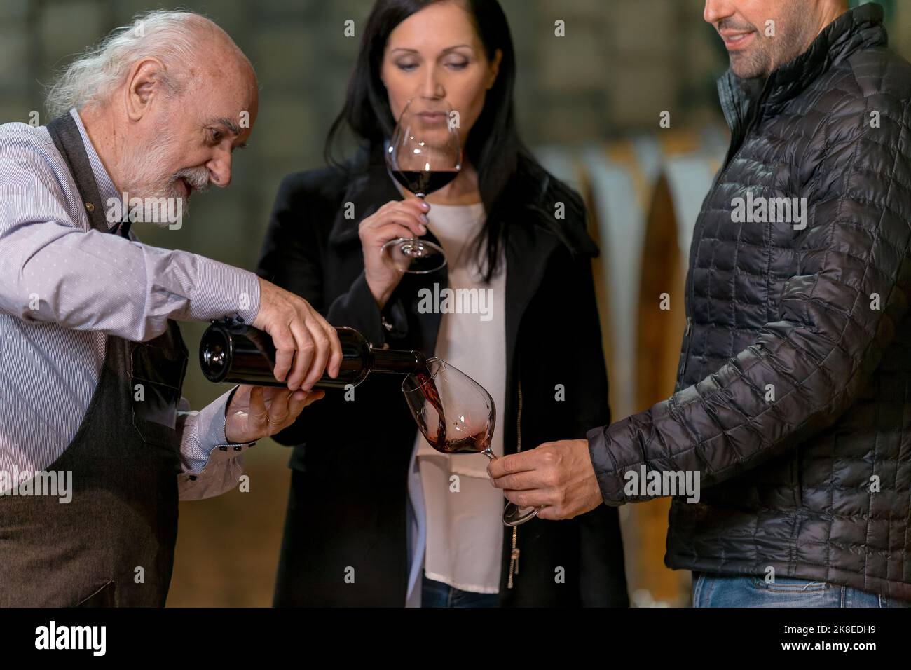 Homme caucasien, un vignetier, parlant et goûtant du vin avec un couple de clients dans la salle de stockage du domaine, dans une cave à vin avec de gros barils Banque D'Images