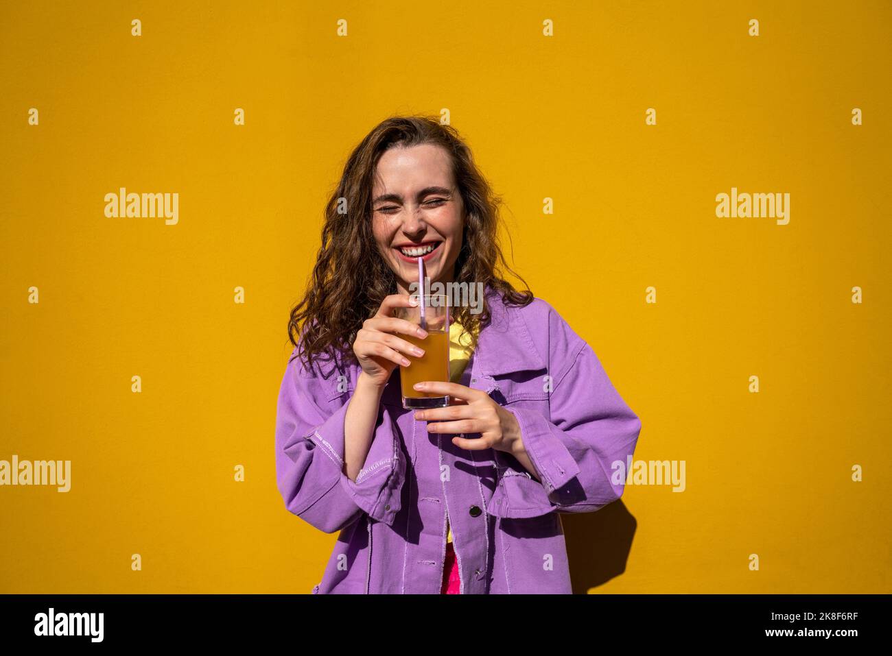 Femme gaie avec les yeux fermés buvant du jus devant le mur jaune Banque D'Images