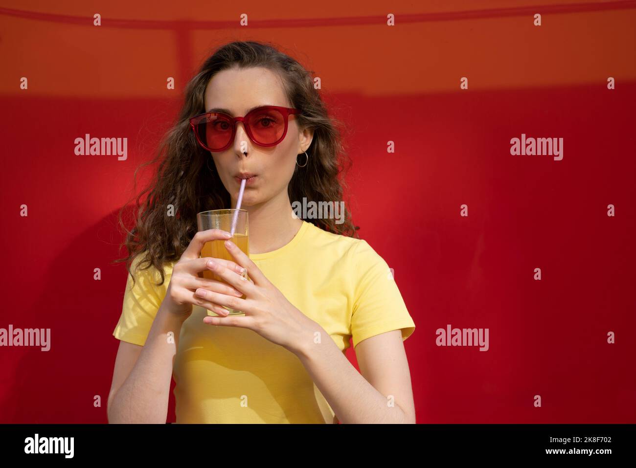 Femme buvant du jus portant des lunettes de soleil devant le mur rouge Banque D'Images