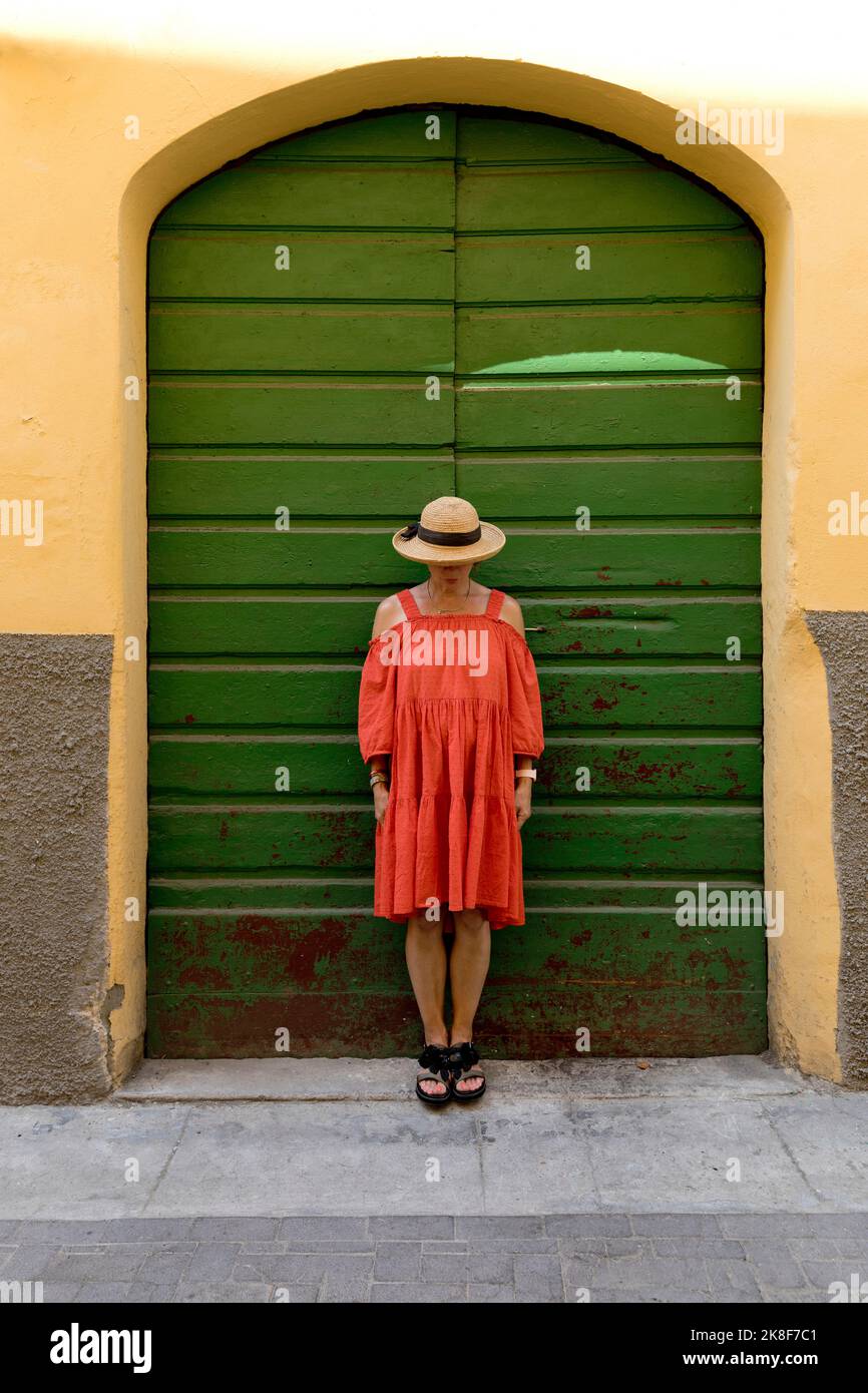 Femme âgée portant une robe rouge et un chapeau de soleil devant la porte verte Banque D'Images