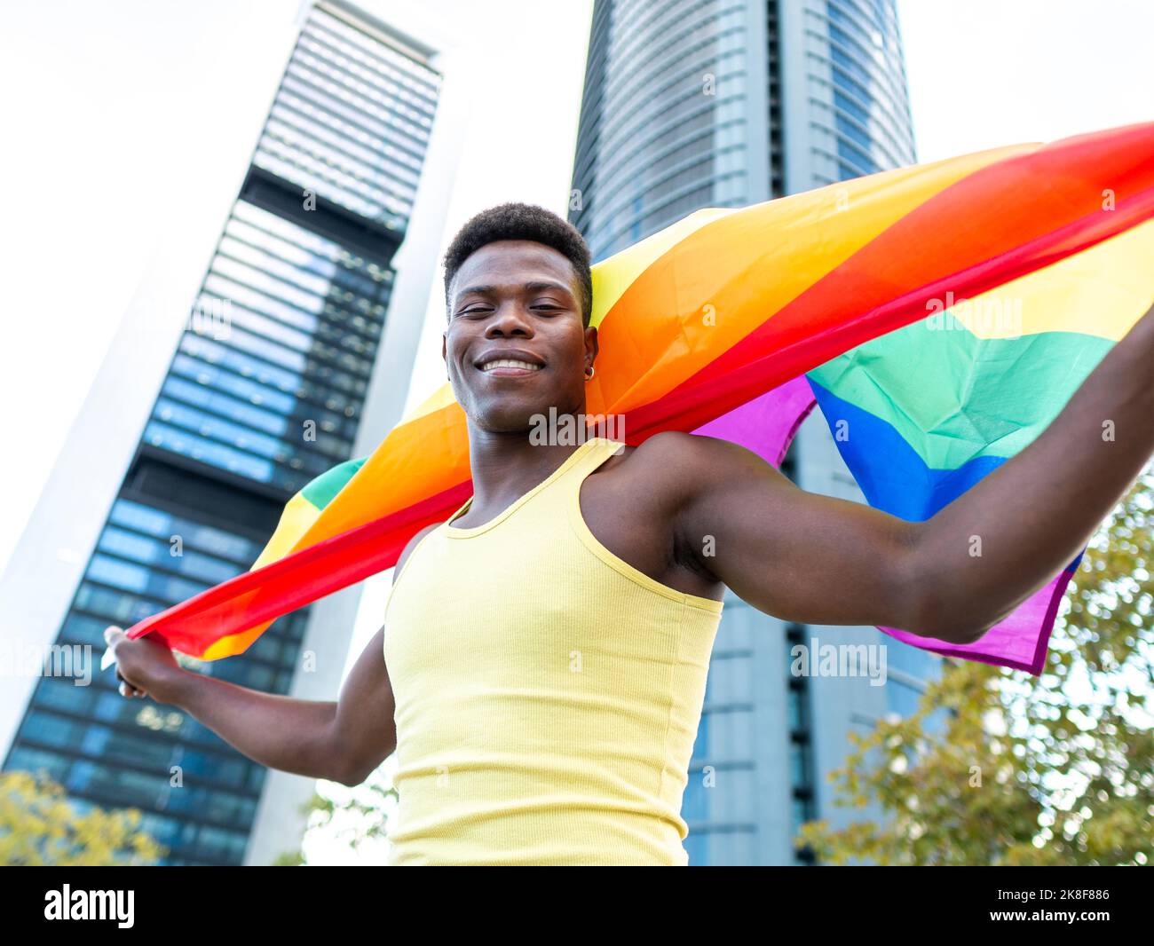 Jeune homme souriant portant une veste jaune portant un drapeau arc-en-ciel devant des bâtiments modernes Banque D'Images