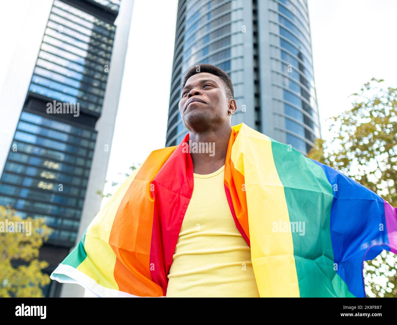 Jeune homme heureux enveloppé dans un drapeau arc-en-ciel devant des gratte-ciel Banque D'Images