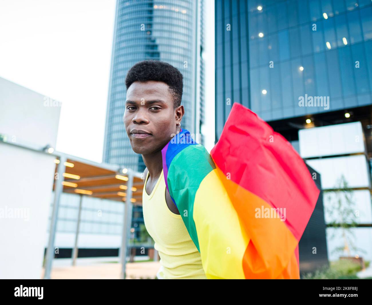 Jeune homme enveloppé dans un drapeau arc-en-ciel par des bâtiments modernes Banque D'Images
