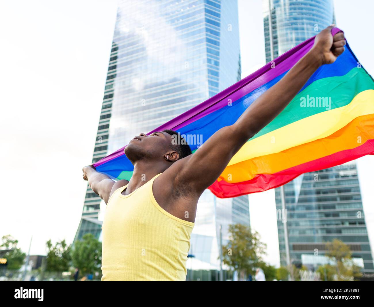 Jeune homme portant une veste jaune portant un drapeau arc-en-ciel devant les gratte-ciel Banque D'Images