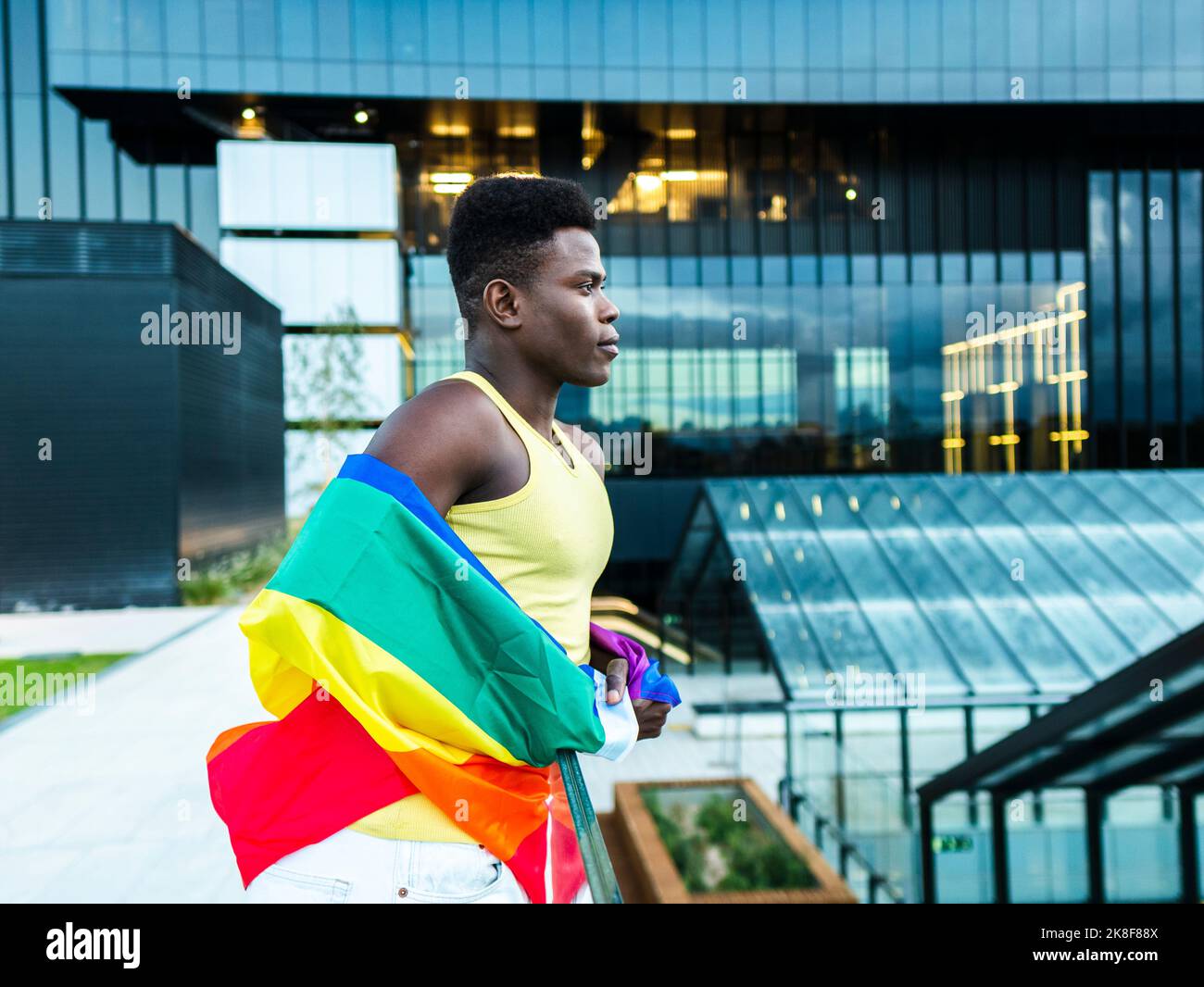Jeune homme confiant enveloppé d'un drapeau arc-en-ciel, appuyé sur une rampe par des bâtiments modernes en verre Banque D'Images