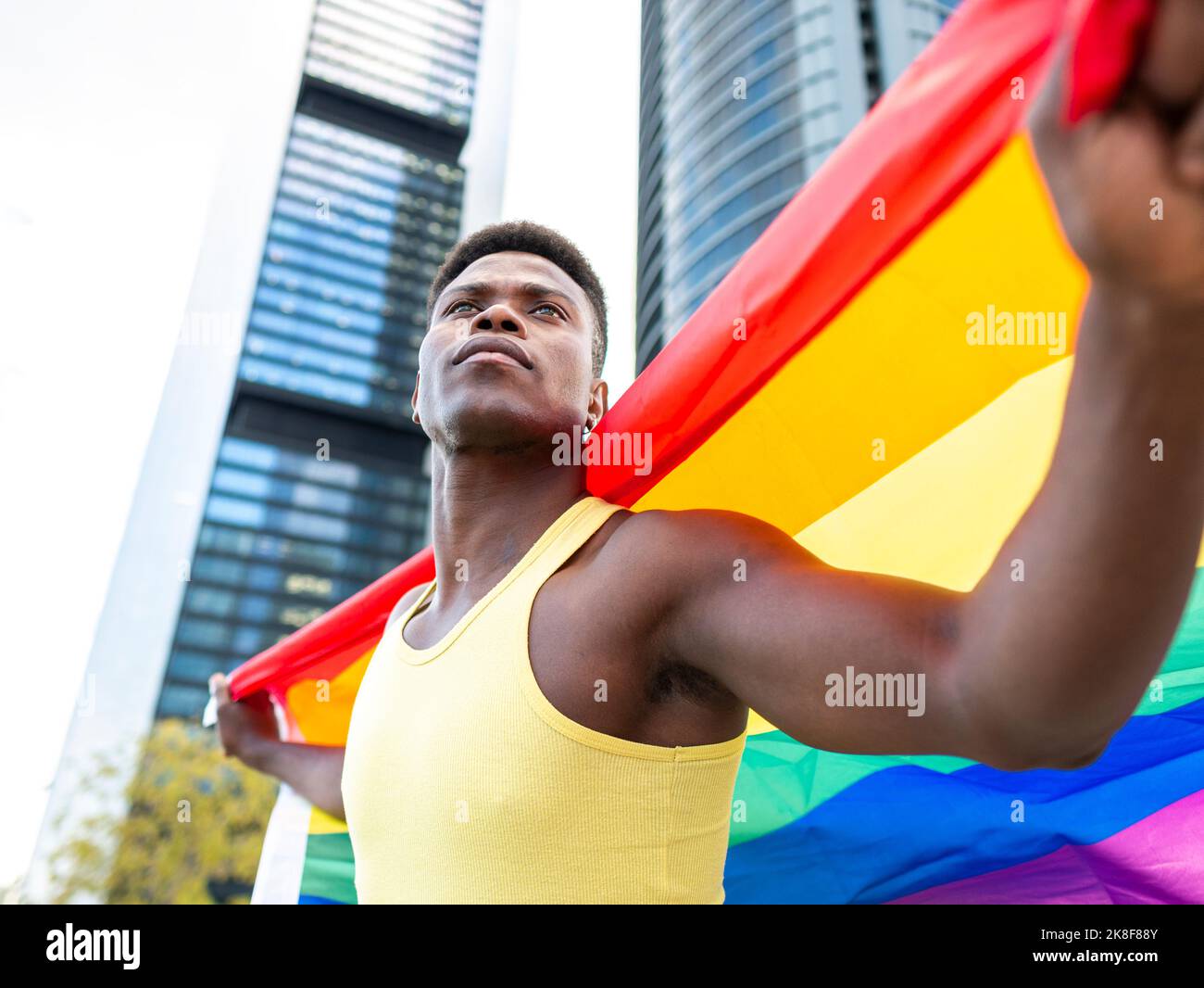 Jeune homme attentionné portant un drapeau arc-en-ciel devant des bâtiments modernes Banque D'Images