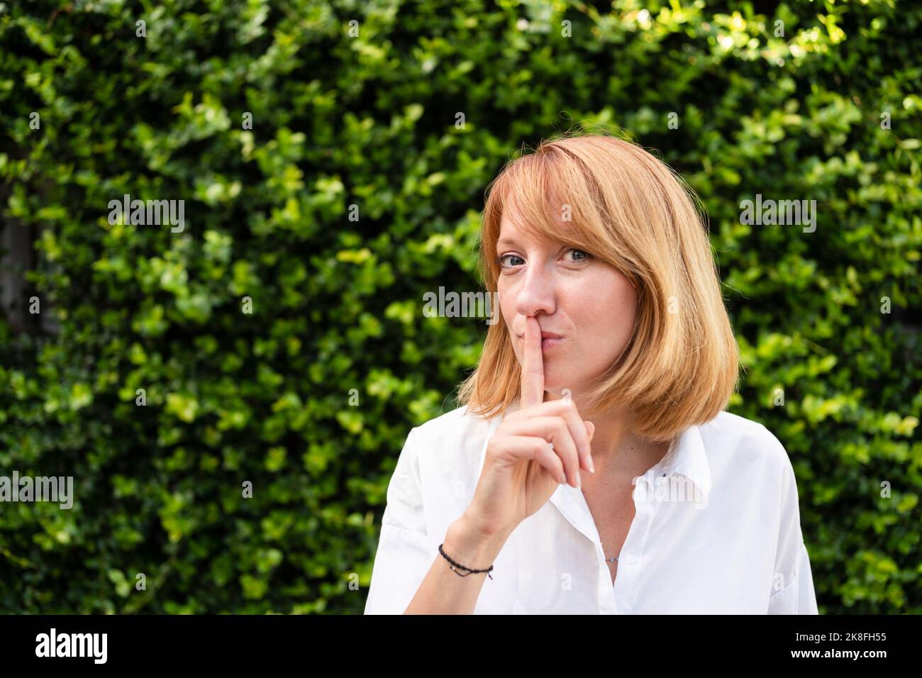 Femme avec le doigt sur les lèvres debout devant la haie verte Banque D'Images