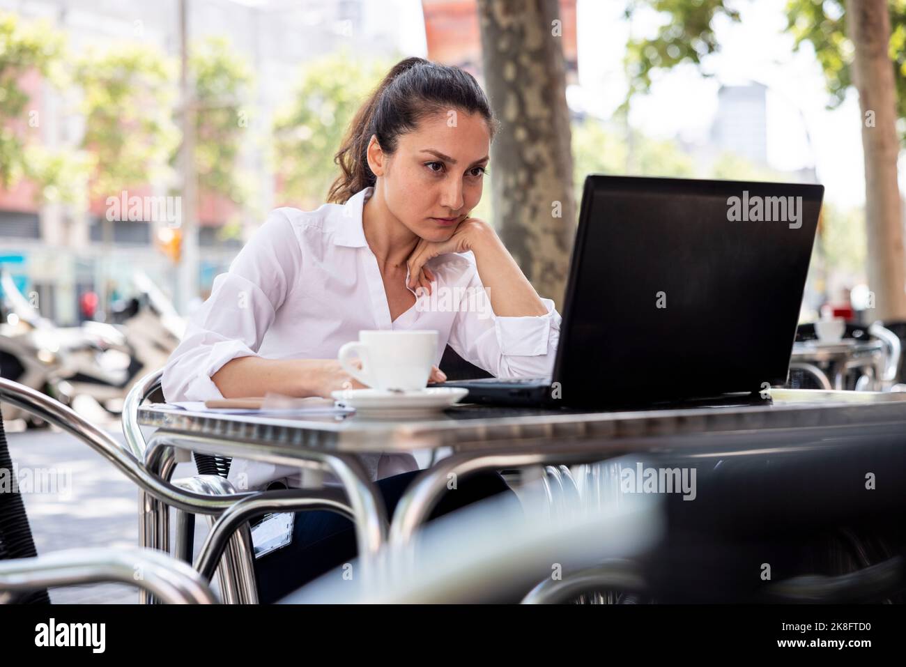 Femme d'affaires avec main sur le menton travaillant sur ordinateur portable au café-terrasse Banque D'Images