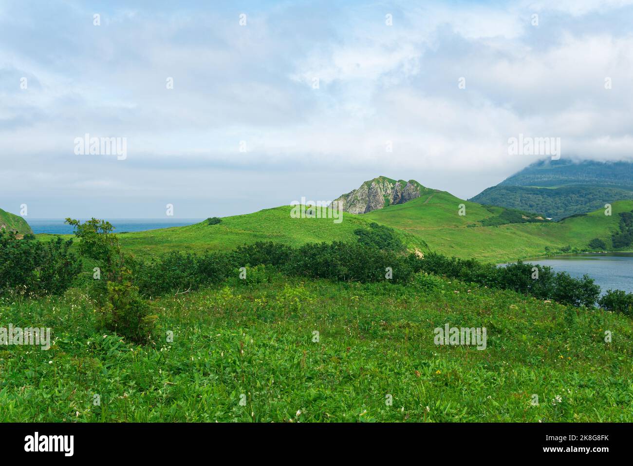 Paysage naturel de l'île de Kunashir avec des collines herbeuses, des roches volcaniques, un volcan dans les nuages et une vallée avec un lagon Banque D'Images