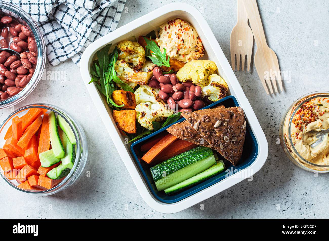 Cuisine vegan panier repas plat. Houmous, haricots, légumes cuits au four, salade verte, pain de seigle et bâtonnets de légumes dans un récipient en plastique, vue du dessus. Banque D'Images