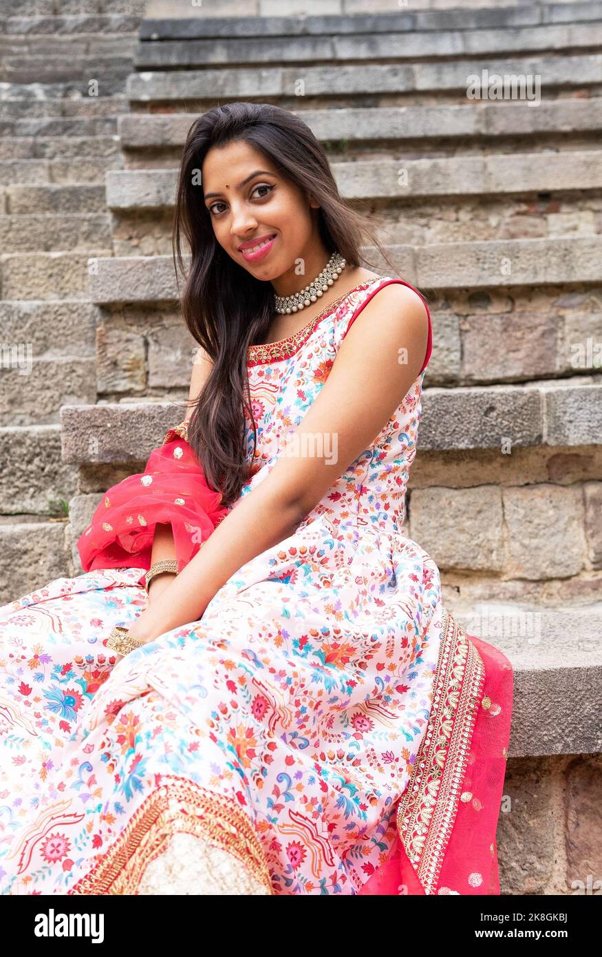 Jeune femme indienne souriante en robe traditionnelle et avec de longs cheveux assis sur les escaliers en pierre et regardant la caméra Banque D'Images