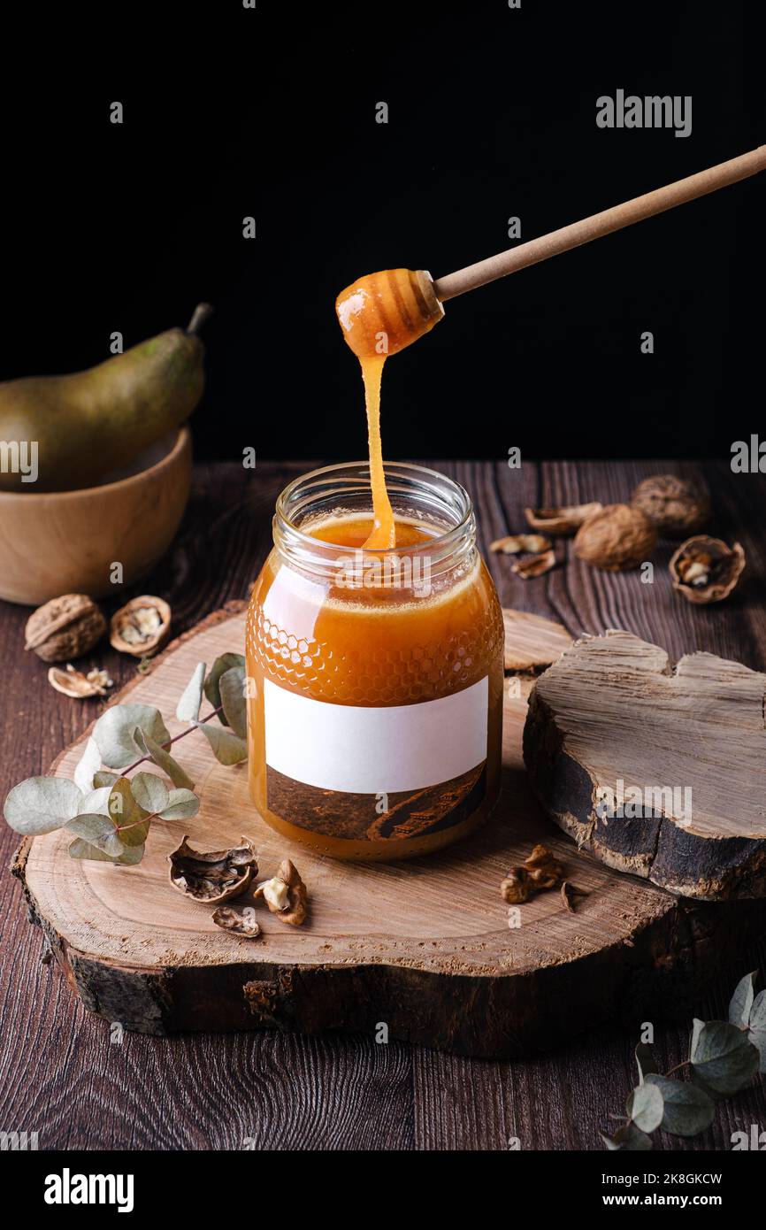Filet de miel naturel à angle élevé s'écoulant du balancier en bois dans un pot en verre avec une étiquette vierge placée sur une dalle de bois près des noix et de la poire contre le noir Banque D'Images