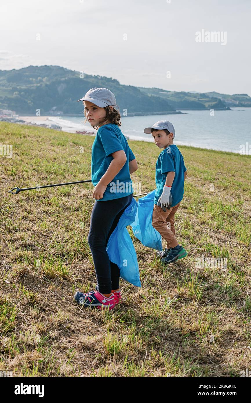 Des enfants écoactivistes dans des vêtements de sport avec des sacs de bac bleu collectant des déchets sur le flanc vert herbacé à flanc de colline près de mer bleu calme le jour ensoleillé Banque D'Images