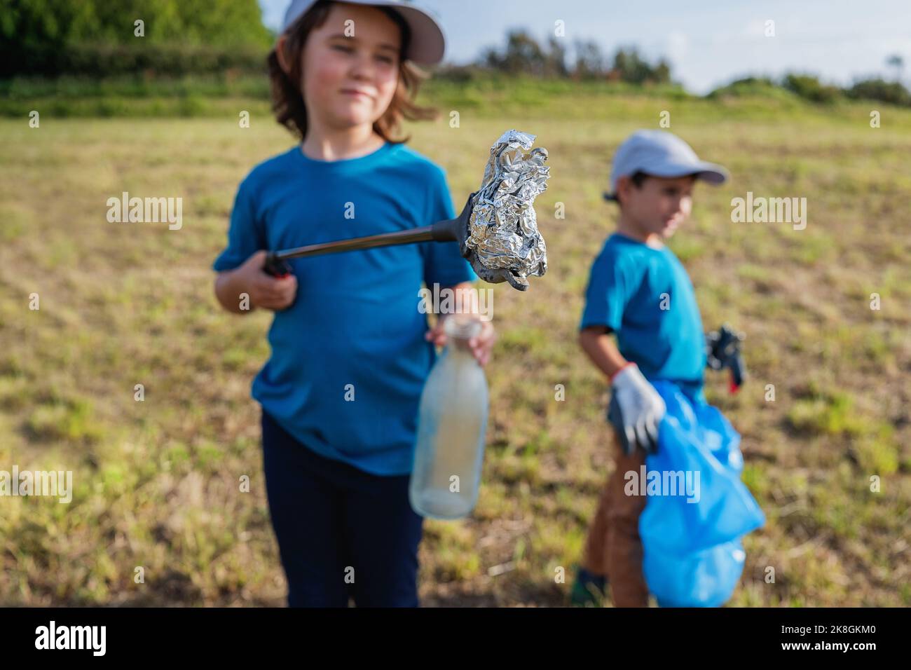 Préadolescents volontaires dans des t-shirts et casquettes bleus debout avec des pinces de thrash et un sac poubelle tout en collectant les déchets en plastique sur la pelouse herbacée Banque D'Images Préadolescents volontaires dans des t-shirts et casquettes bleus debout avec des pinces de thrash et un sac poubelle tout en collectant les déchets en plastique sur la pelouse herbacée Banque D'Images