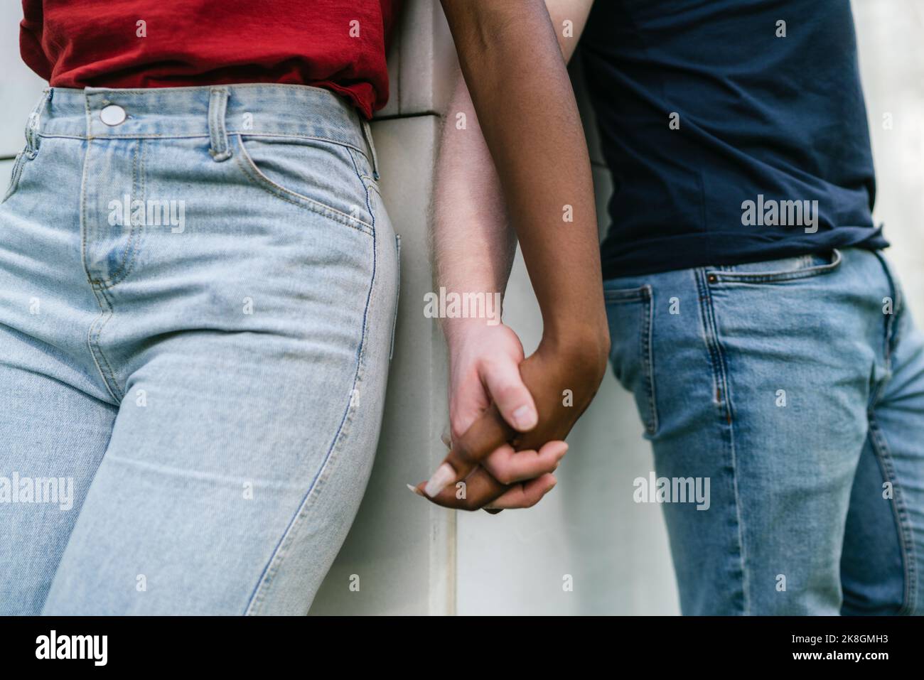 Cachez un petit ami et une petite amie multiracial méconnaissable en jeans et t-shirts inclinés sur un mur blanc et tenant les mains Banque D'Images Cachez un petit ami et une petite amie multiracial méconnaissable en jeans et t-shirts inclinés sur un mur blanc et tenant les mains Banque D'Images