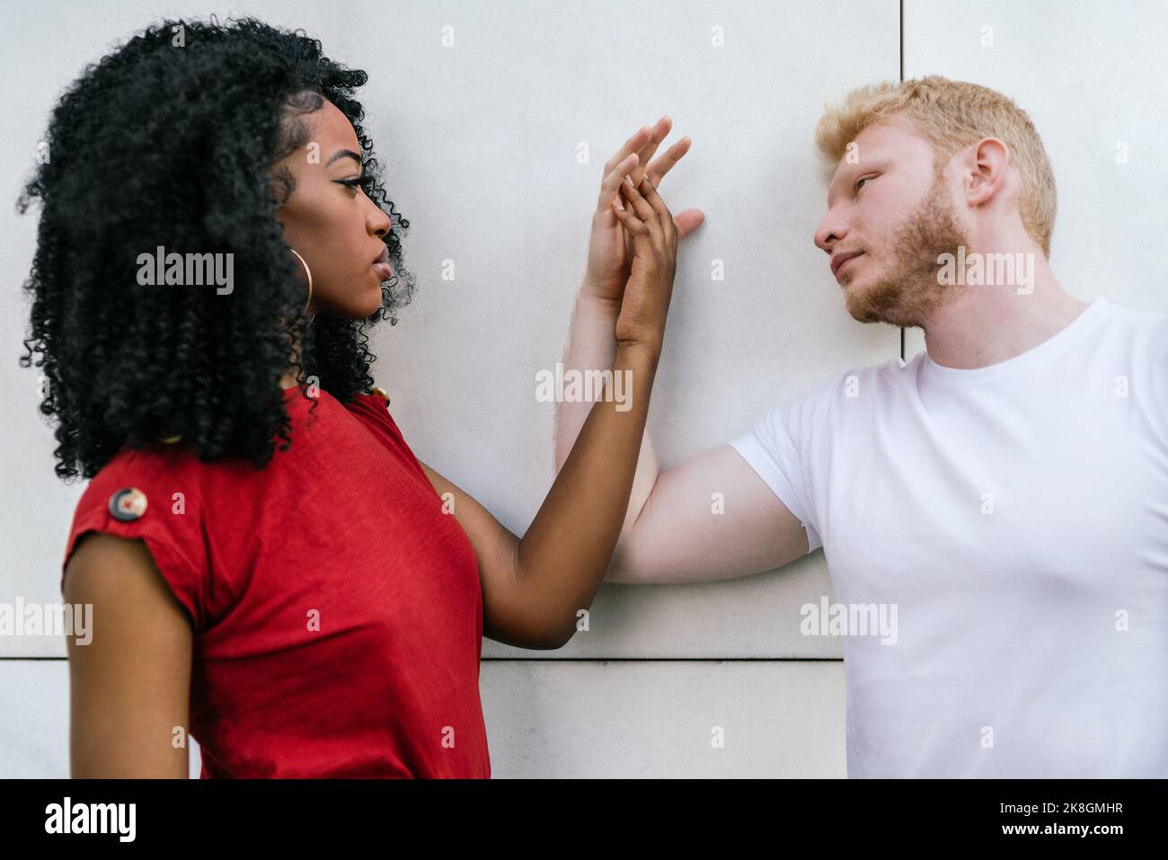 Vue latérale de la petite amie afro-américaine et de l'ami blond dans les t-shirts pentus sur le mur de carrelage blanc et touchant les mains tout en regardant chaque o Banque D'Images Vue latérale de la petite amie afro-américaine et de l'ami blond dans les t-shirts pentus sur le mur de carrelage blanc et touchant les mains tout en regardant chaque o Banque D'Images