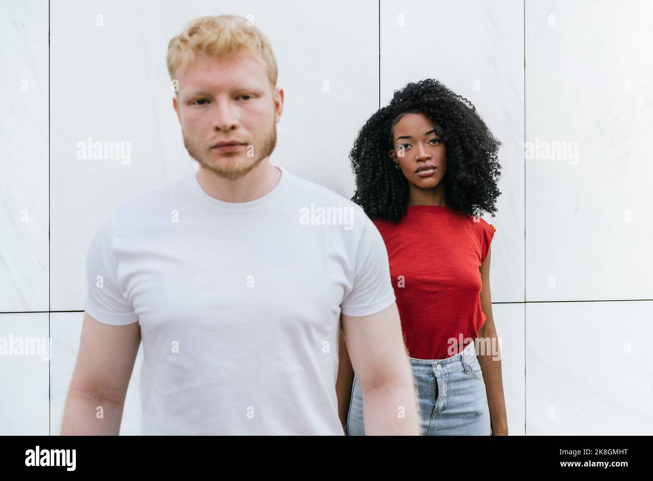 Femme afro-américaine sans émotion avec maquillage debout sur le mur blanc du bâtiment près de l'homme blond barbu dans le t-shirt blanc tout en regardant la caméra Banque D'Images