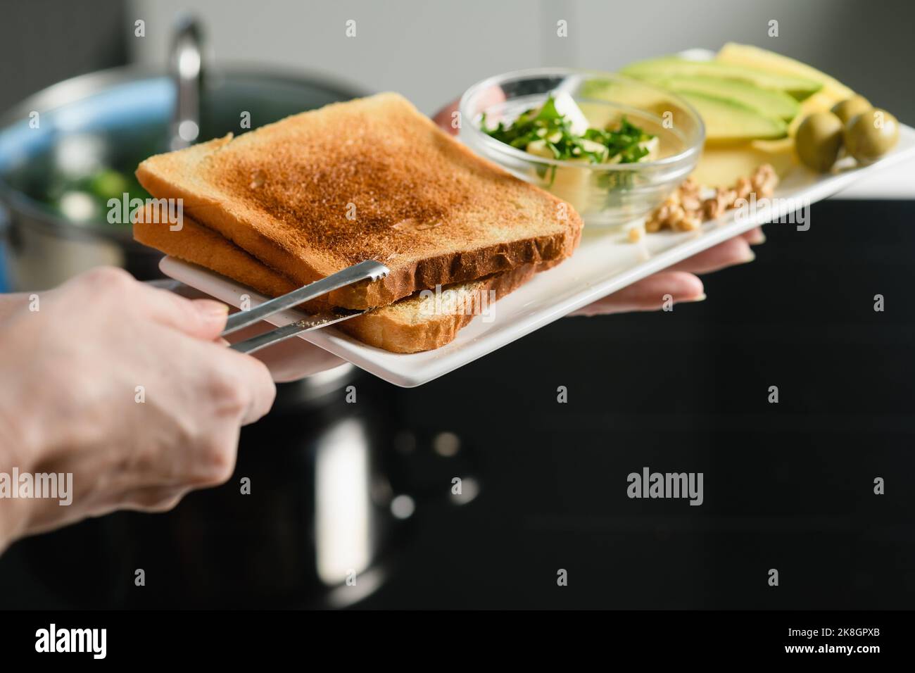 La main de femme met du pain grillé fraîchement cuit sur une assiette. Tranches d'avocat, beurre, fromage pour un sandwich sain. Banque D'Images