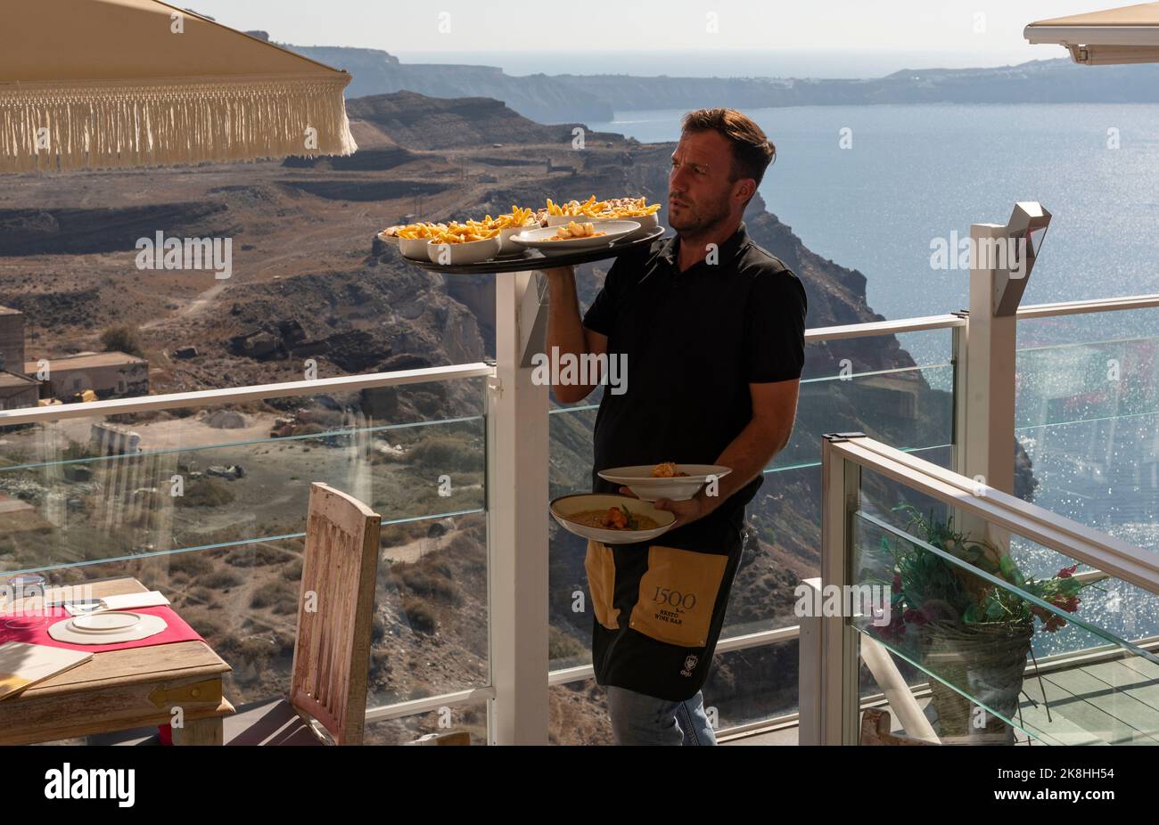 Fira, Santorin, Grèce. 2022. Un serveur transportant des plats sur un bistro de restaurant à Fira, Santorini surplombant la mer Egée, Santorini. Banque D'Images