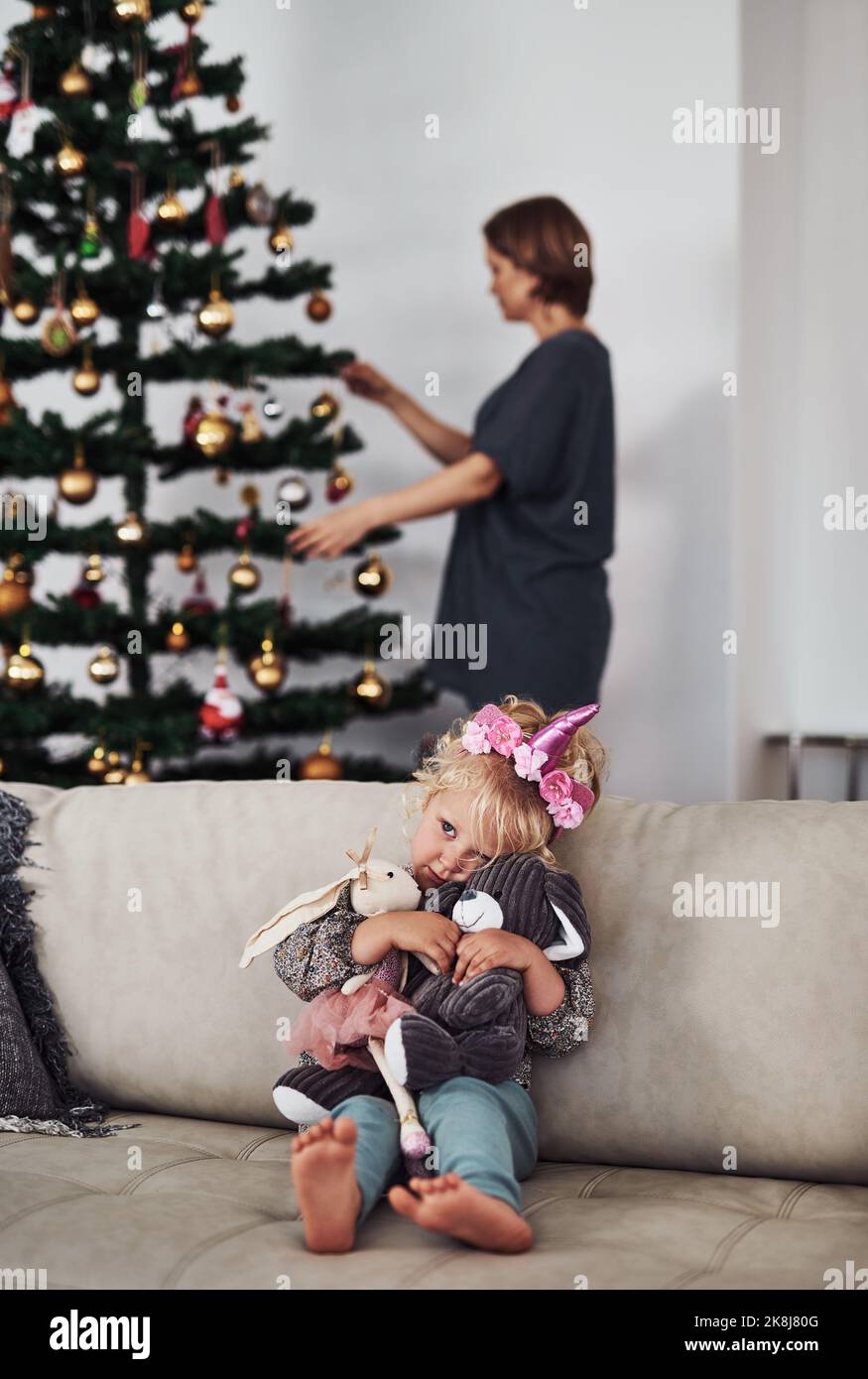 Peut-on jouer maintenant maman. Une adorable petite fille assise sur le canapé tandis que sa mère fixe l'arbre de Noël derrière elle. Banque D'Images