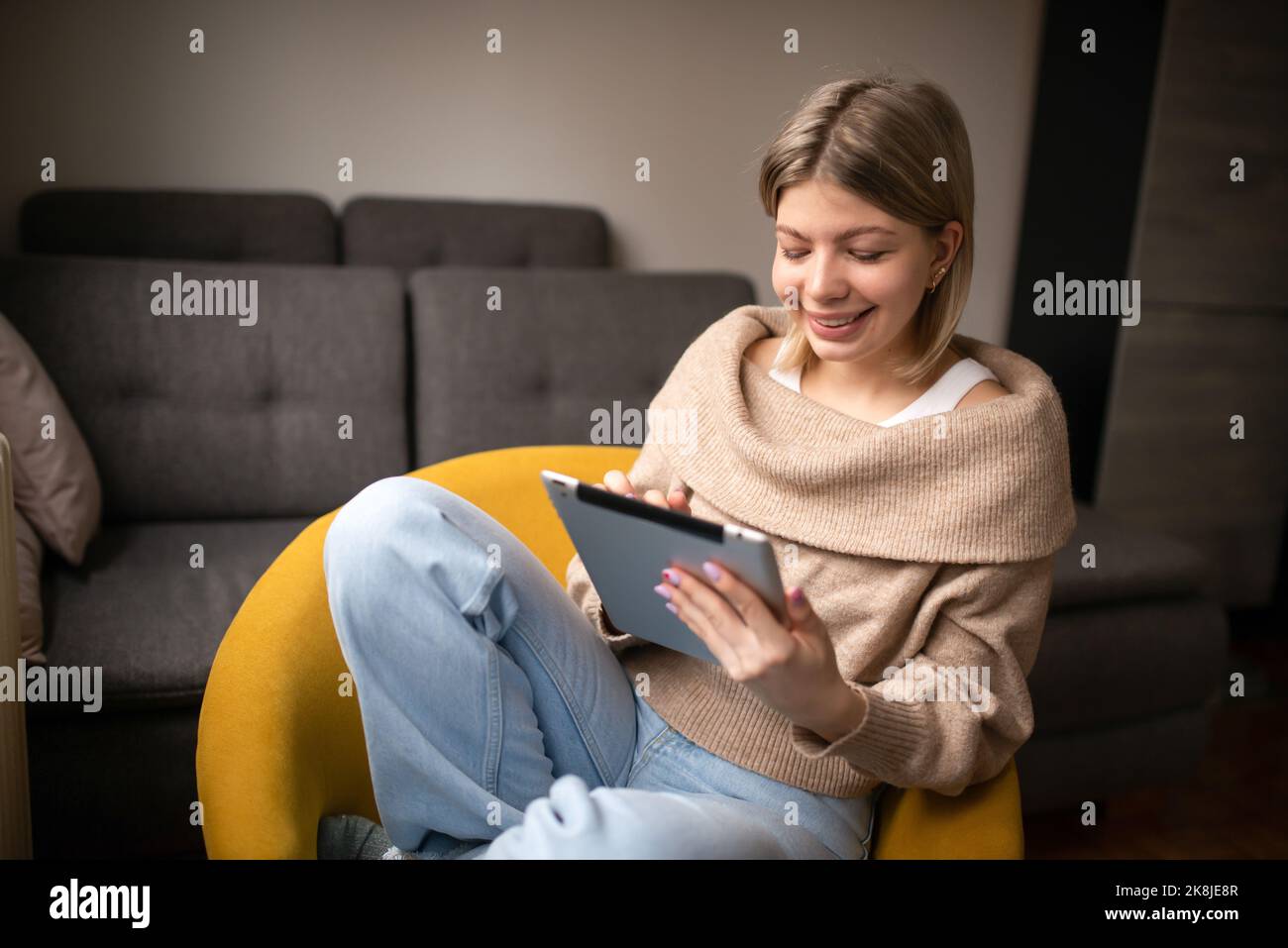 Une femme heureuse et belle qui regarde des vidéos ou qui profite de contenus de divertissement sur une tablette, assise sur le canapé de la maison. Banque D'Images