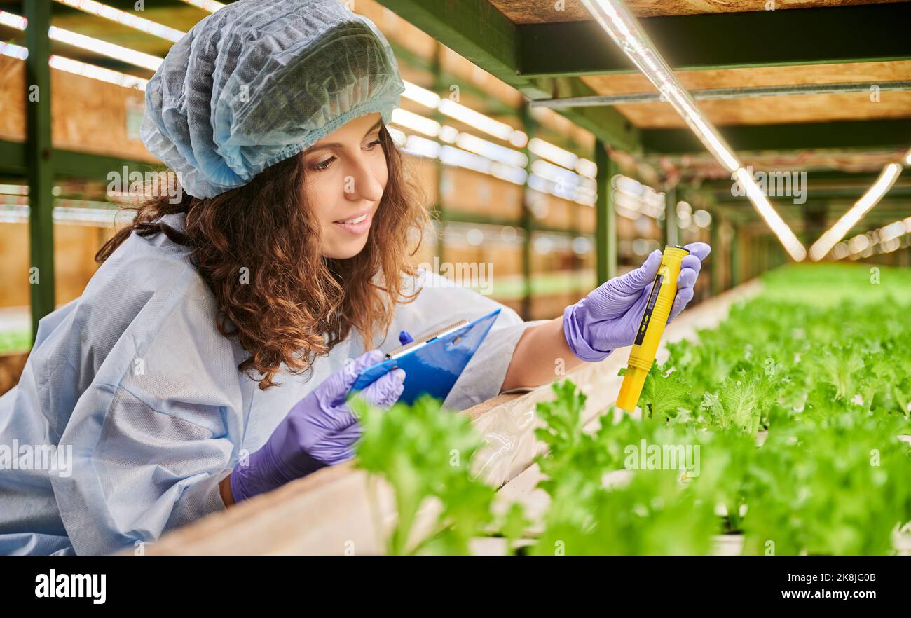 Femme agronome utilisant un pH-mètre en serre. Une jardinière féminine heureuse tient le presse-papiers tout en mesurant la valeur du pH du sol avec un appareil électronique numérique. Banque D'Images