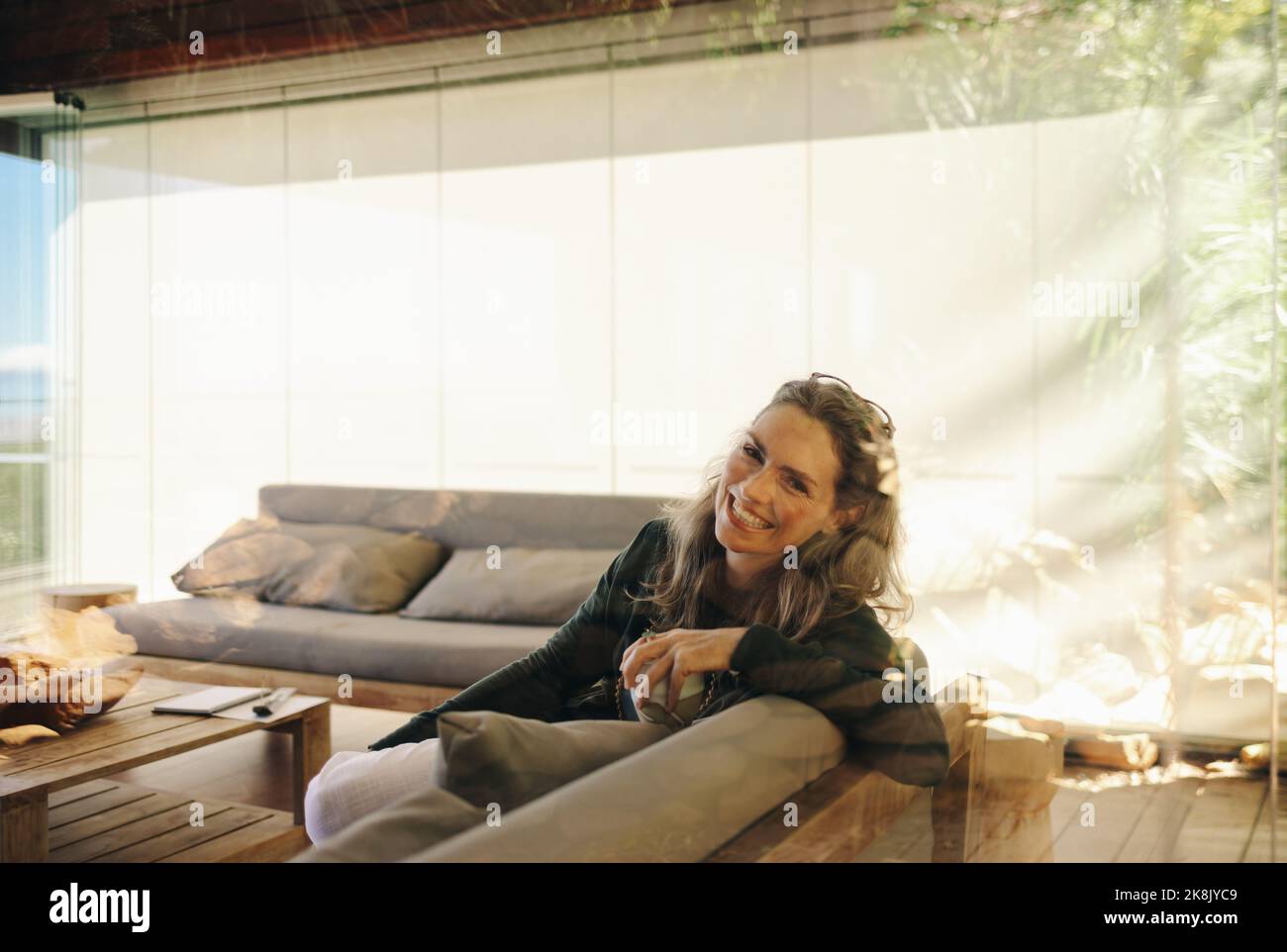 Bonne femme mûre souriant à la caméra tout en étant assise sur un canapé avec une tasse de thé. Une femme âgée et gaie se détendant dans son salon à la maison. Banque D'Images
